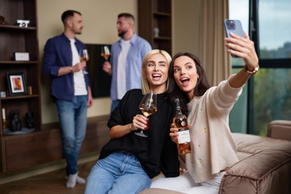 Doua femei isi fac selfie cu  vin rose, intr-o sufragerie